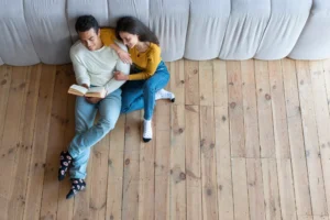 Top-down view of a happy couple sitting on a wooden floor leaning against a pale gray sofa